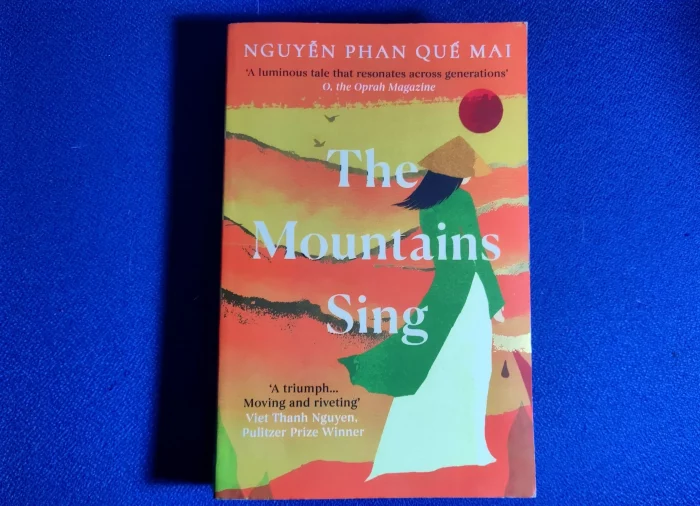 Sur un tissu bleu, un livre orange est déposé. Son titre est The Mountains Sing. De couleur dominante orange, il représente une femme portant un chapeau de paille qui tourne le dos au spectateur et observe des montagnes.