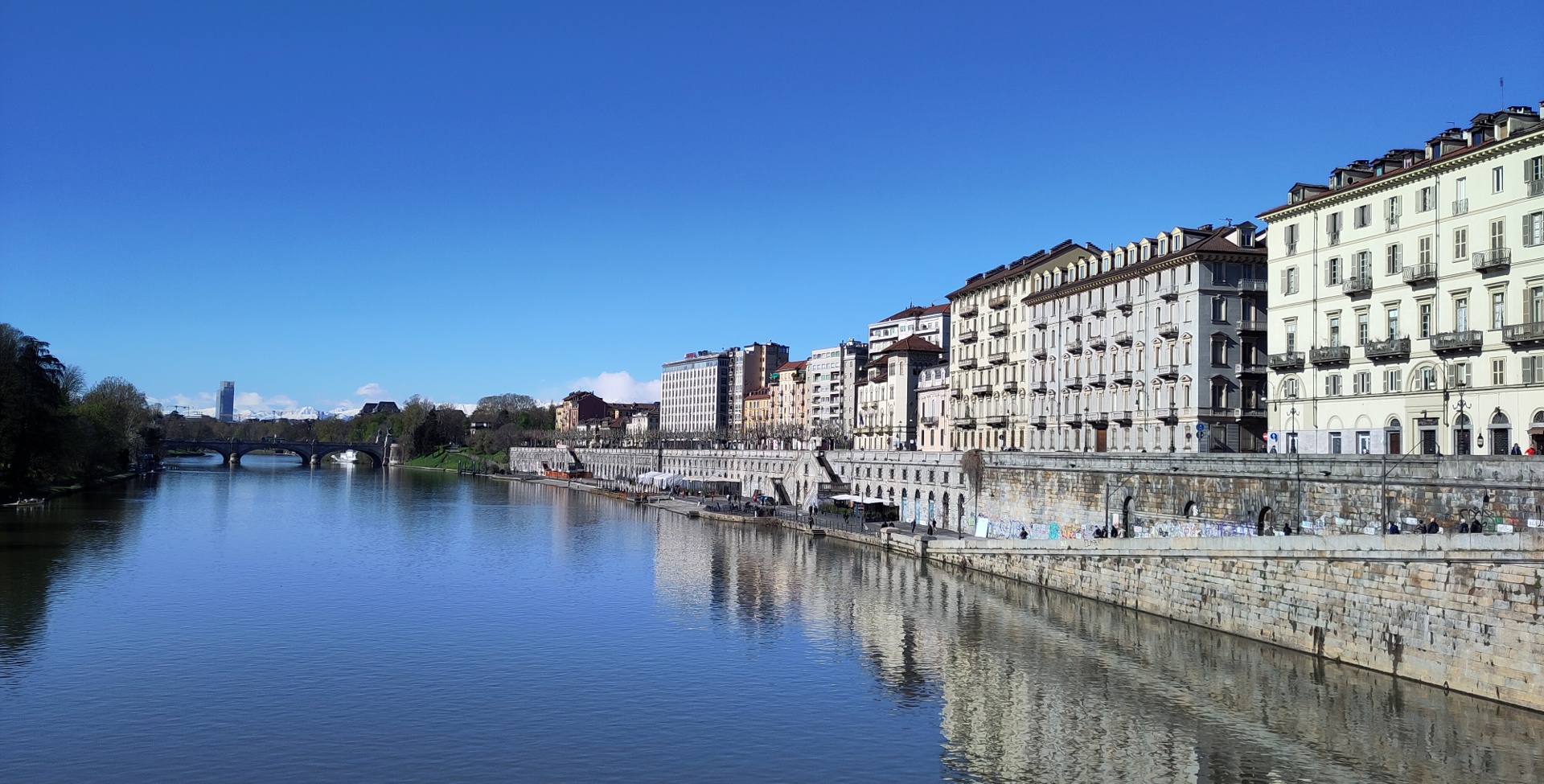 A sunny view of the Torino quays, with buildings on the right. Very far away, you can see snowy mountaintops.