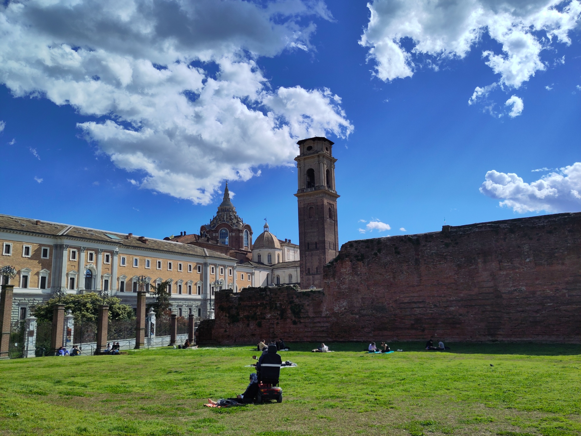 An extremely sunny park. A couple, a man in a wheelchair and a woman wearing a hijab, is enjoying the sun on the grass. Further out, the Palatine door, a tower and a church.