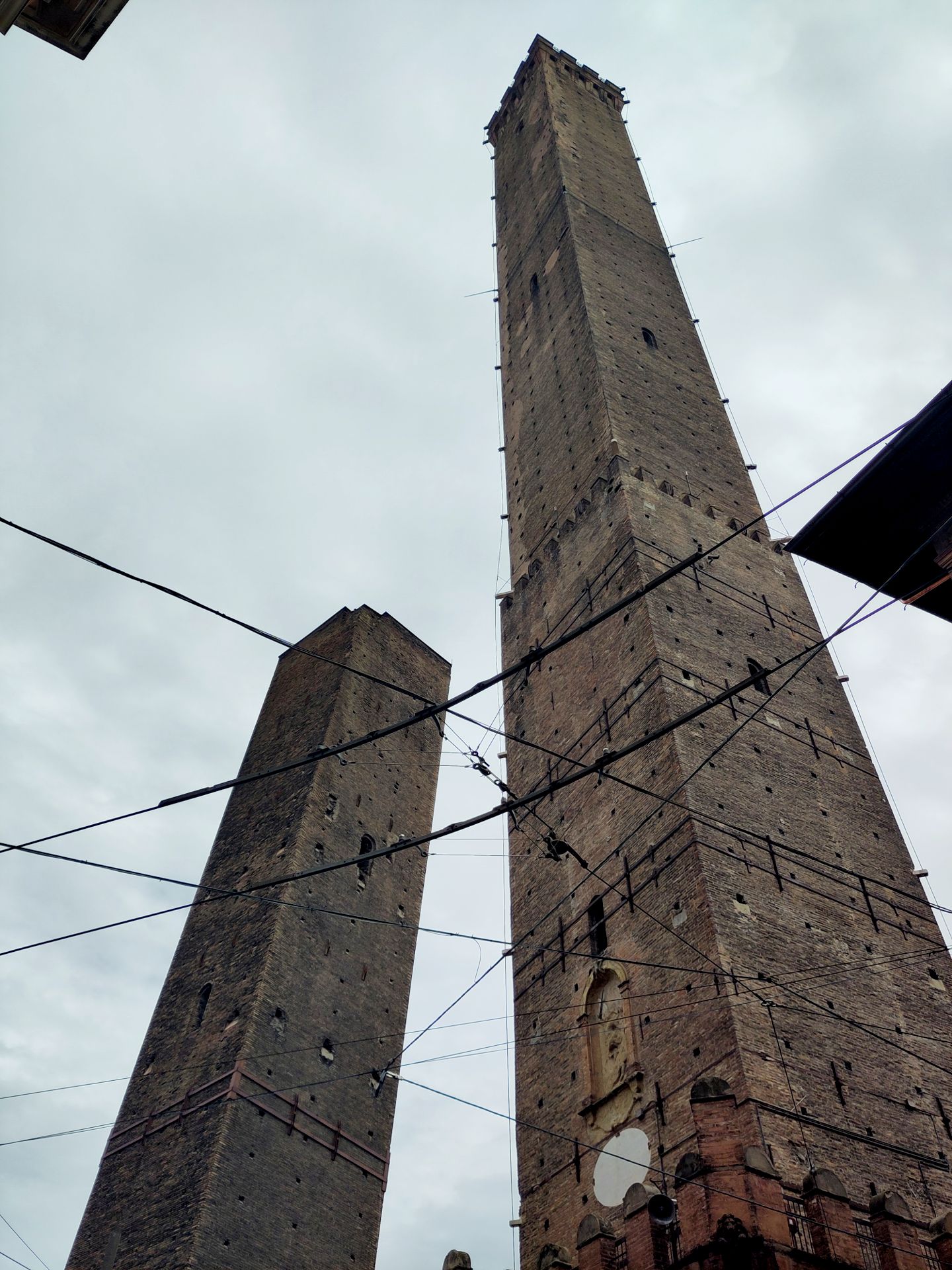 Two grey and not very vertical towers made of stones. In front of them, many interweaving electric cables for the tramway. It's cloudy so all is different shades of grey.