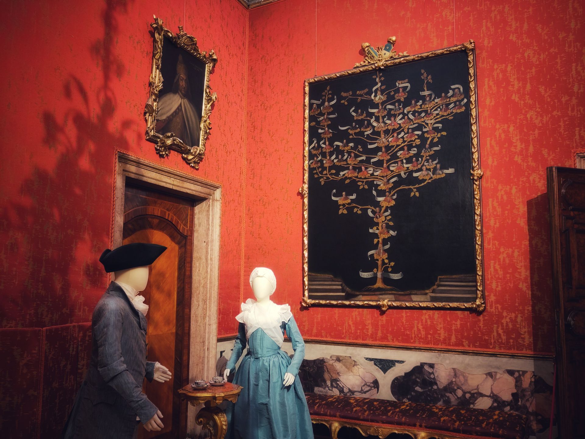 A couple of mannequins, dressed as Venetian nobles from the 18th century. The walls are very red and there's an old-school family tree on one wall.