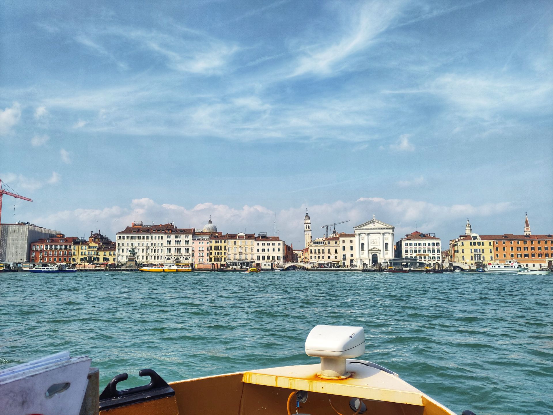 A view of the Venice shore, including yellow and orange buildings and a church belltower. I'm on a boat, so the front of the boat is visible right in front of me.