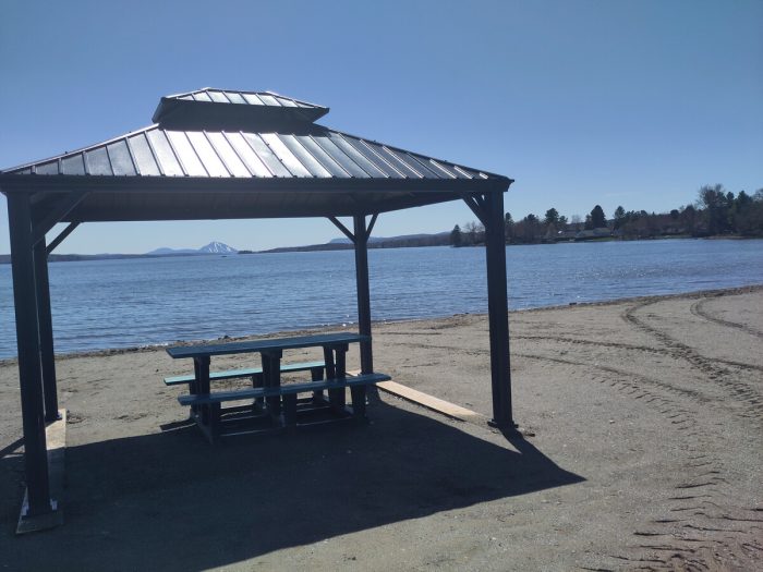 A beautiful sunny day. On a beach, there's a picnic table under a little roof. Far away at the other side of the lake, a mountain.